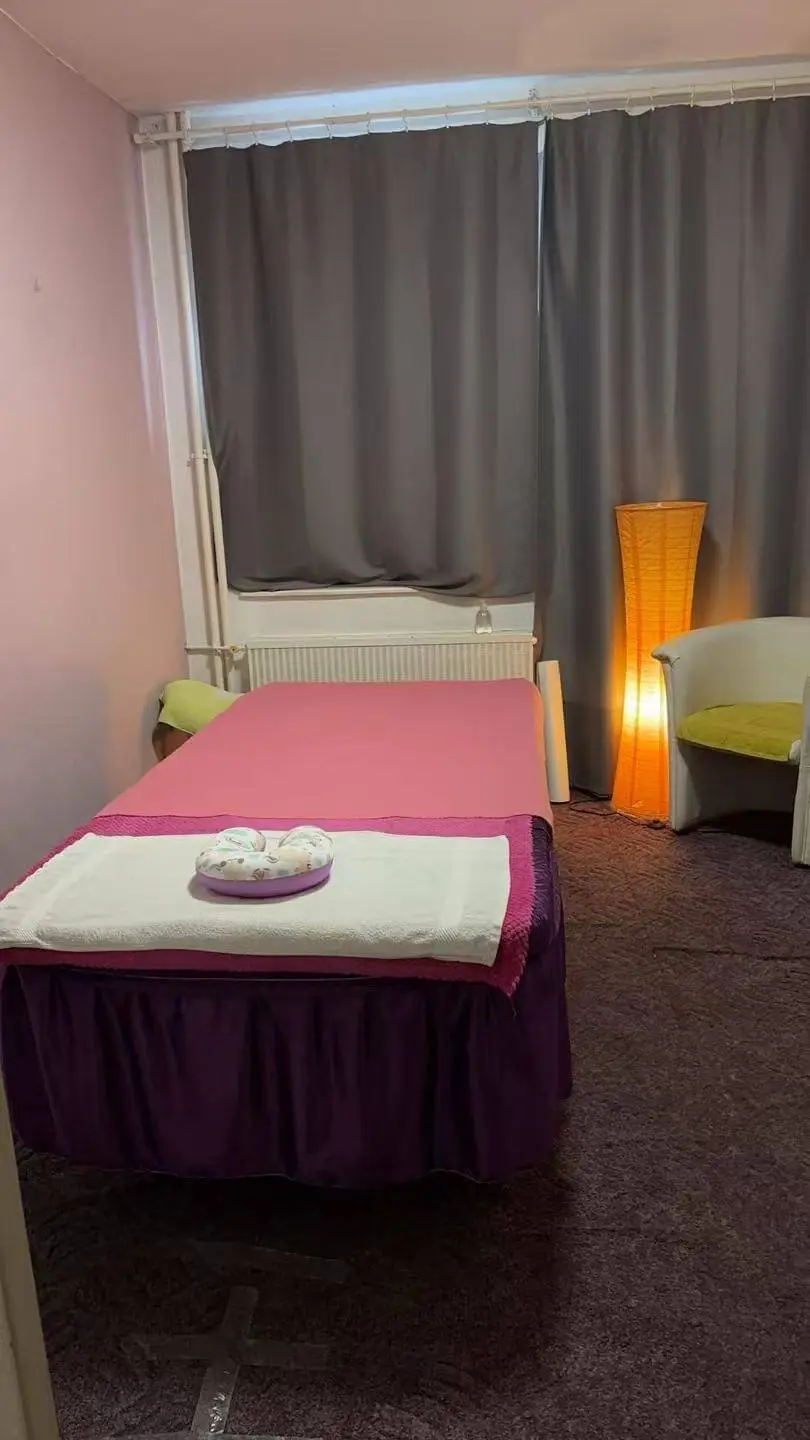 Massageraum mit orange-violettem Massagebett auf gemustertem Teppich, cremefarbenen Wänden und Blumendekoration.